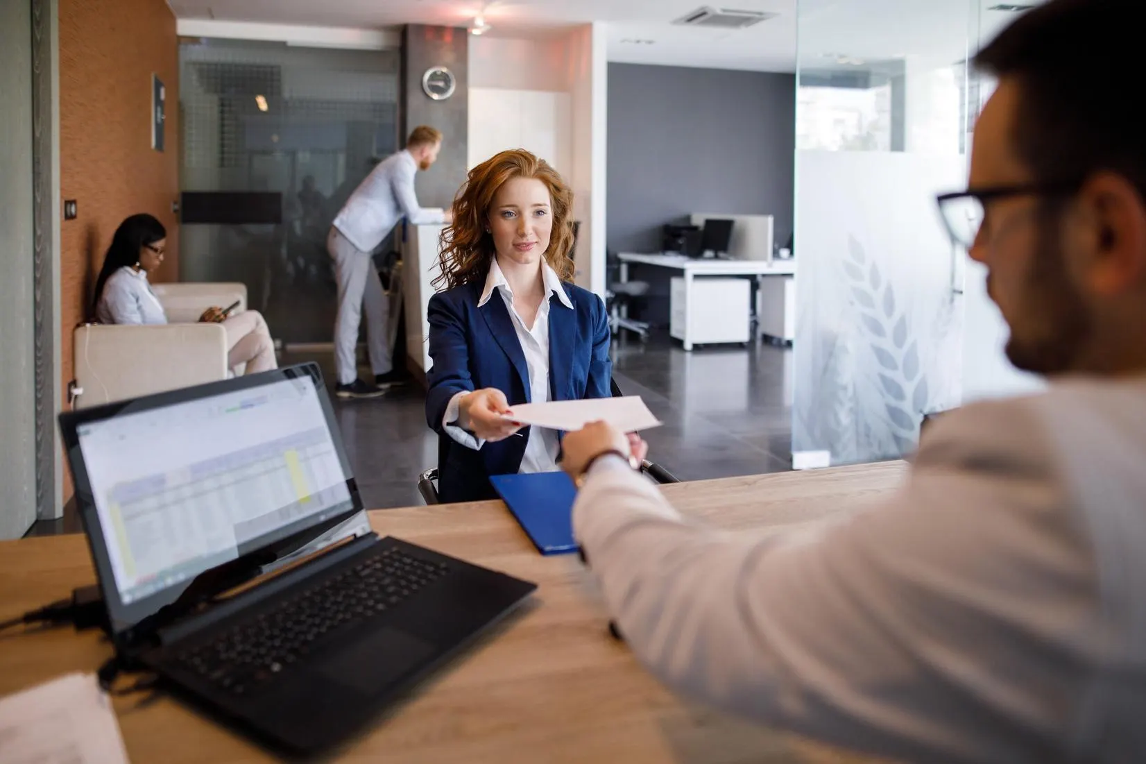 A woman in a blue suit hands over a document to a man at a desk.